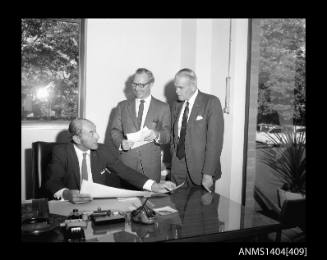 Photographic negative showing a group of three men looking at plans on an office desk