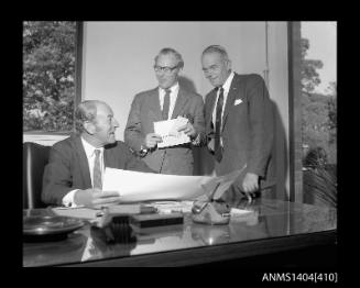 Photographic negative showing a group of three men looking at plans on an office desk