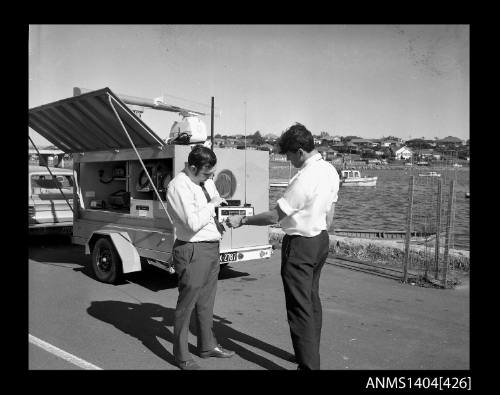 Photographic negative showing two men demonstrating navigational equipment contained in an AWA marine service trailer