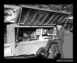 Photographic negative showing two men demonstrating navigational equipment contained in an AWA marine service trailer