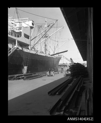 Photographic negative showing building materials about to be loaded onto a vessel, with Glebe Island Bridge in the background