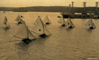 Postcard featuring a photograph of nine skiff sailing in the water, with land in the background, and a large vessel near the skiffs on the right hand side of the photograph