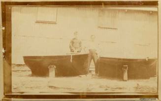 Black and white photograph of two men standing beside two small boats