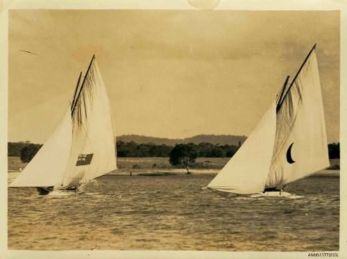 Black and white photograph of two skiffs sailing side by side, with land visible behind them