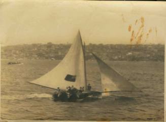 Skiff sailing in the water, with several crew members, and a flag on the sail