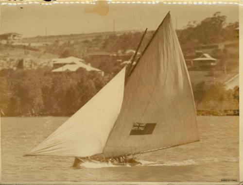 Skiff sailing close to the shoreline, with a Union Jack flag on the sail