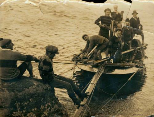 Black and white photograph of two men sitting on a rock, and six men standing in a small boat, with the mast resting horizontally on the frame of the boat