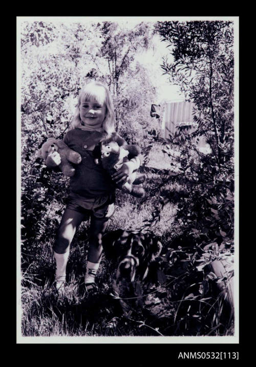 A young girl holding a monkey toy facing the camera with washing on the line