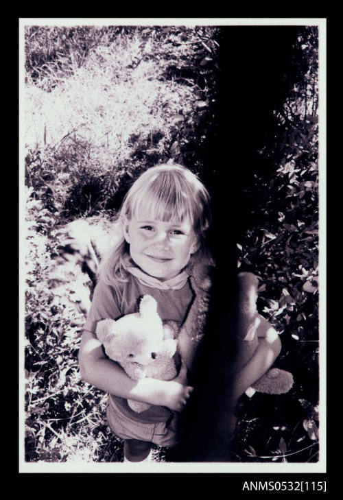 Girl looking dirctly towards the camera while clutching a teddy bear and doll