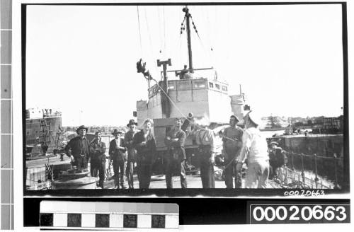 Group of ratings and civilian men on the deck of a naval vessel watching a monkey climb a line