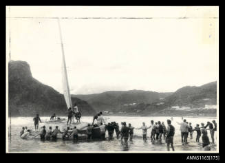 The single masted trimaran with cabin being steadied and towed ashore by about 30 people