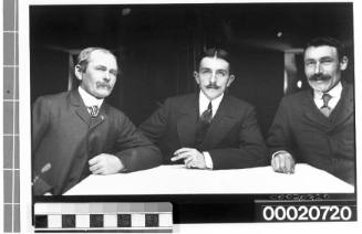 Group portrait of three men in suits and seated at a table