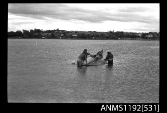 Black and white negative number 3 depicting view of hydroplane stationary in the water with three men knee deep making adjustments