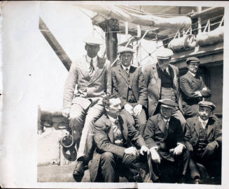 Group of seven men on a wharf in front of the companion way of a vessel with furled sails