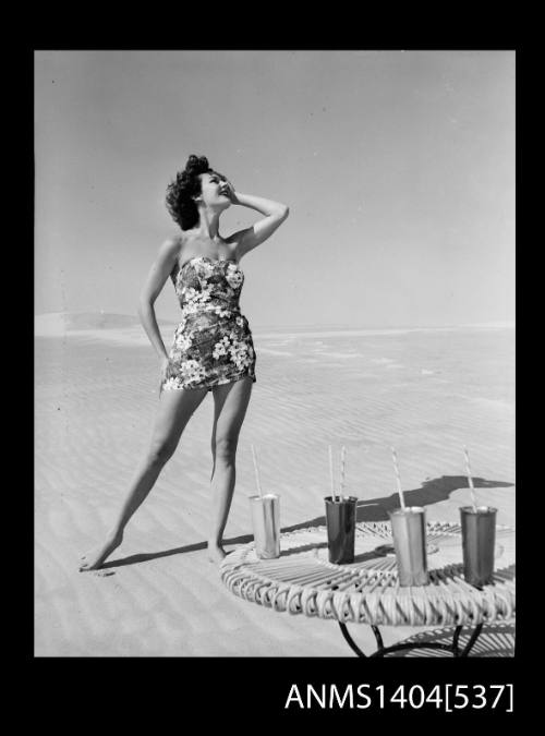 Photographic negative of a swimsuit model posing on a sand dune near a table with drinks