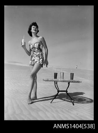 Photographic negative of a swimsuit model posing on a sand dune near a table with drinks