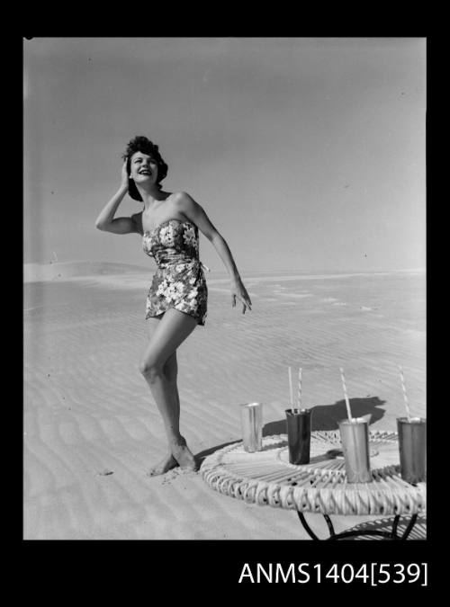 Photographic negative of a swimsuit model posing on a sand dune near a table with drinks