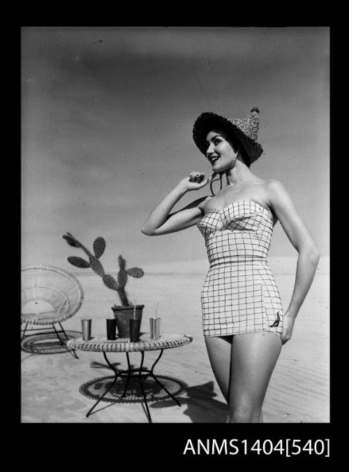 Photographic negative of a swimsuit model posing on a sand dune near a chair and table with drinks