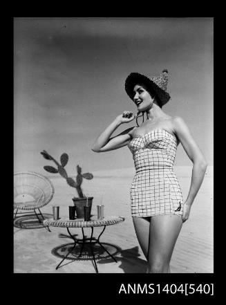 Photographic negative of a swimsuit model posing on a sand dune near a chair and table with drinks