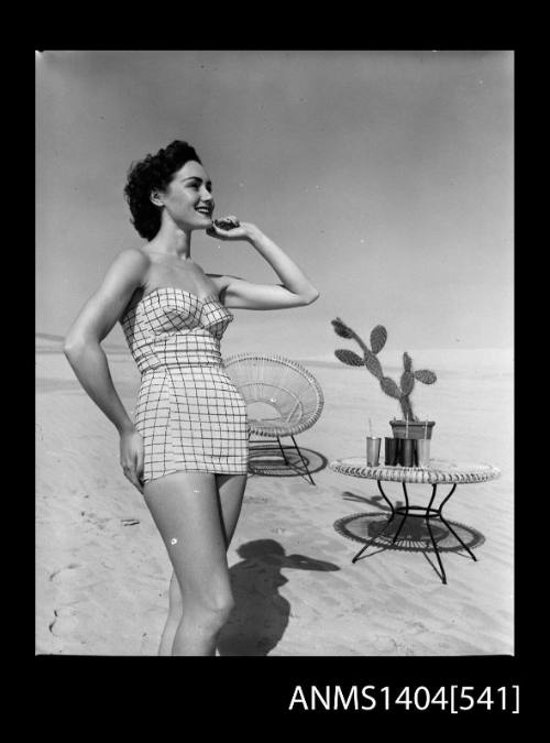 Photographic negative of a swimsuit model posing on a sand dune near a chair and table with drinks