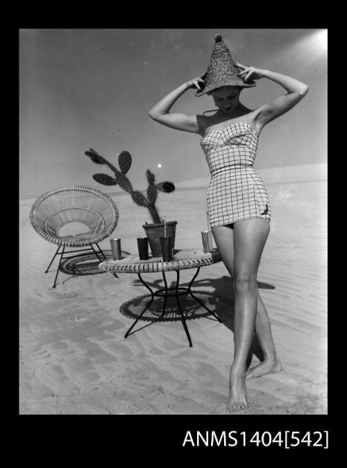 Photographic negative of a swimsuit model posing on a sand dune near a chair and table with drinks