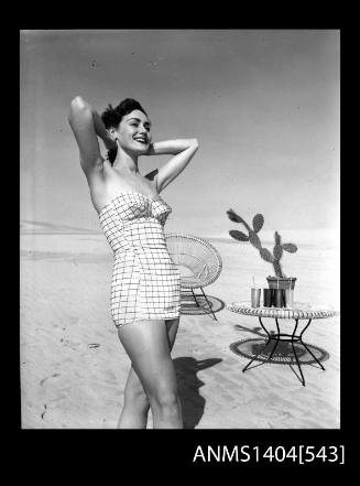 Photographic negative of a swimsuit model posing on a sand dune near a chair and table with drinks