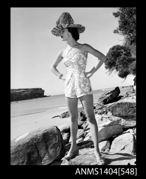 Photographic negative of a swimsuit model posing with a hat on rocks by a seashore