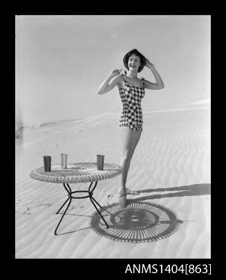 Photographic negative of a swimsuit model posing on a sand dune near a table with drinks