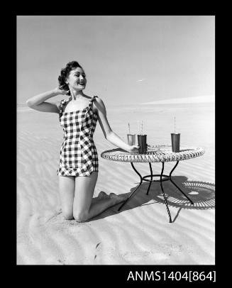 Photographic negative of a swimsuit model posing on a sand dune near a table with drinks