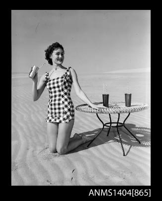 Photographic negative of a swimsuit model posing on a sand dune near a table with drinks