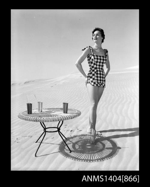 Photographic negative of a swimsuit model posing on a sand dune near a table with drinks