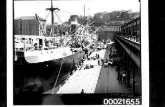 Wharf labourers using derricks to load bales of wool and crates into the holds of SS CANADIAN CRUISER 