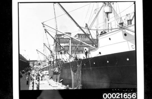 Wharf labourers using derricks to load bales of wool and crates into the holds of SS CANADIAN CRUISER 