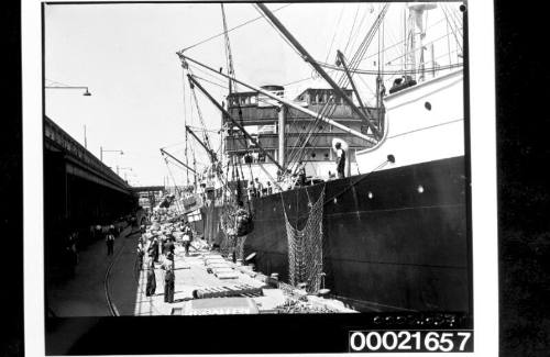 Wharf labourers using derricks to load bales of wool and crates into the holds of SS CANADIAN CRUISER 