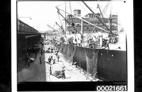 Wharf labourers using derricks to load bales of wool and crates into the holds of SS CANADIAN CRUISER 