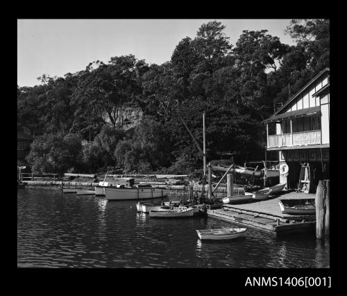 Negative depicting several moored boats, a small jetty and a boat shed with trees and a cliff in the background