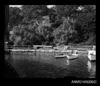 Negative depicting three moored boats, a sandstone retaining wall and wooden fence with trees in the background and a car on the river bank