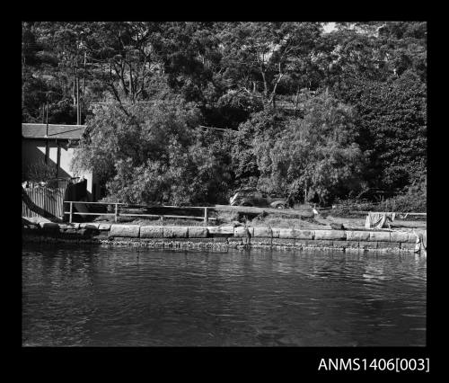 Negative depicting a car on a river bank with buildings to the left and a sandstone retaining wall