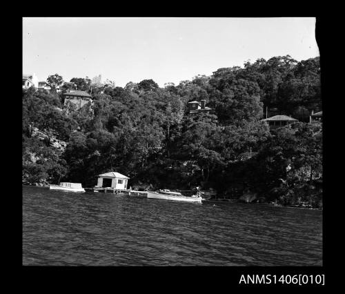 Negative depicting two white boats on the river, near a boat house, with several homes built on the hill behind