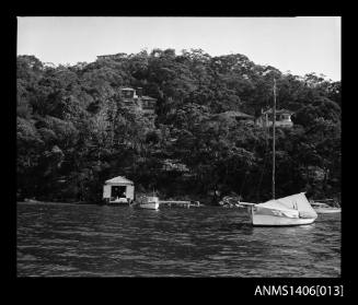 Negative depicting three boats near a shore and a boat house with several homes built on the hill behind