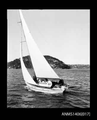 Negative depicting a yacht underway, from portside stern, with a five person crew and headlands with houses are visible in the background