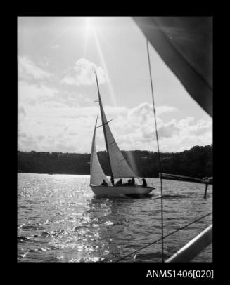 Negative depicting a yacht underway, from portside bow, with a six person crew and headlands with houses are visible in the background