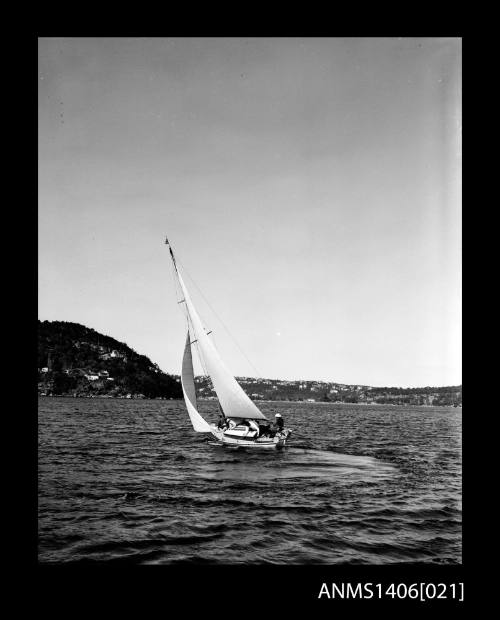 Negative depicting a yacht underway, from portside stern, with a four person crew and headlands with houses are visible in the background