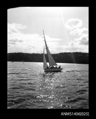 Negative depicting a yacht underway, from portside stern, with a four person crew and headlands with houses are visible in the background