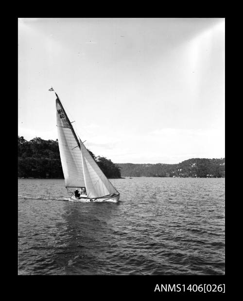 Negative depicting a yacht underway, from starboard bow, with two people visible in the cockpit and headlands with houses are visible in the background