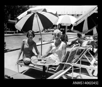 Negative depicting two women in bathing suits, sitting in beach lounge chairs beside a pool