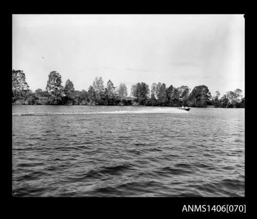 Negative depicting a motorboat at speed, on a river lined with trees on the opposite bank