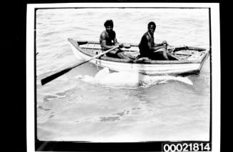 Two Aboriginal men with scarification in rowing boat with a dugong tied to the side of the boat
