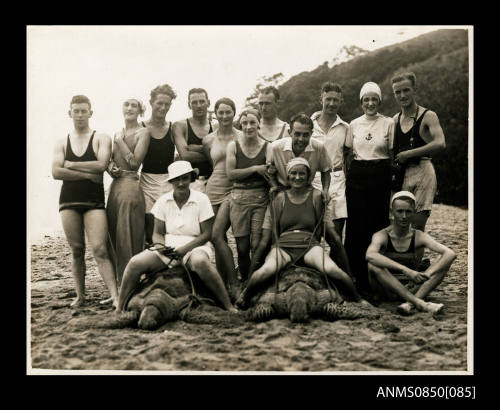 A group of tourists on a sandy beach, including two women seated on turtles