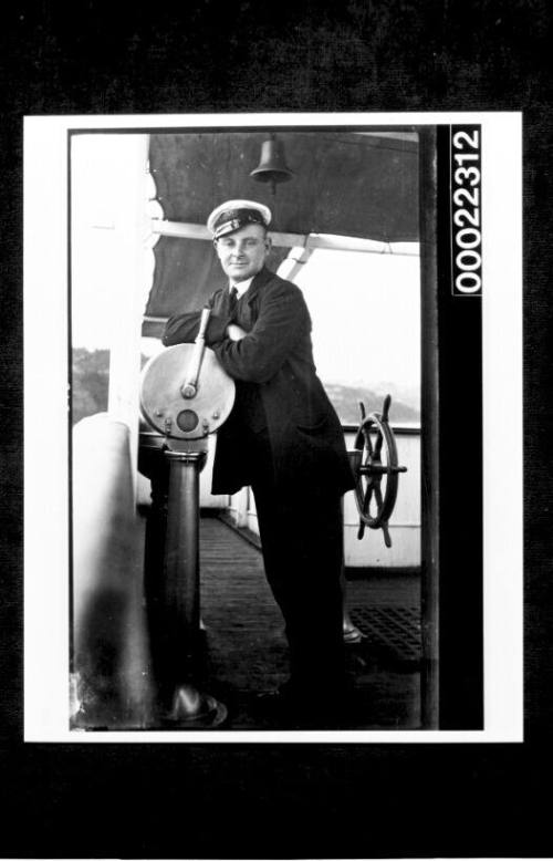 Man in a black suit and naval cap, leaning onto throttle with the ships wheel in the background